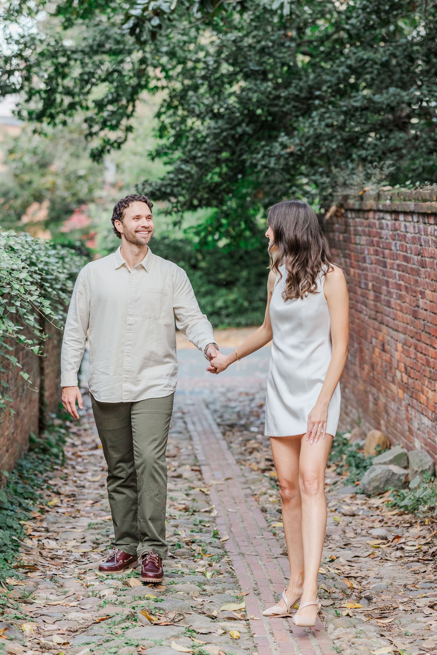 A couple walks hand in hand along a brick path surrounded by lush greenery and a brick wall on a sunny day.