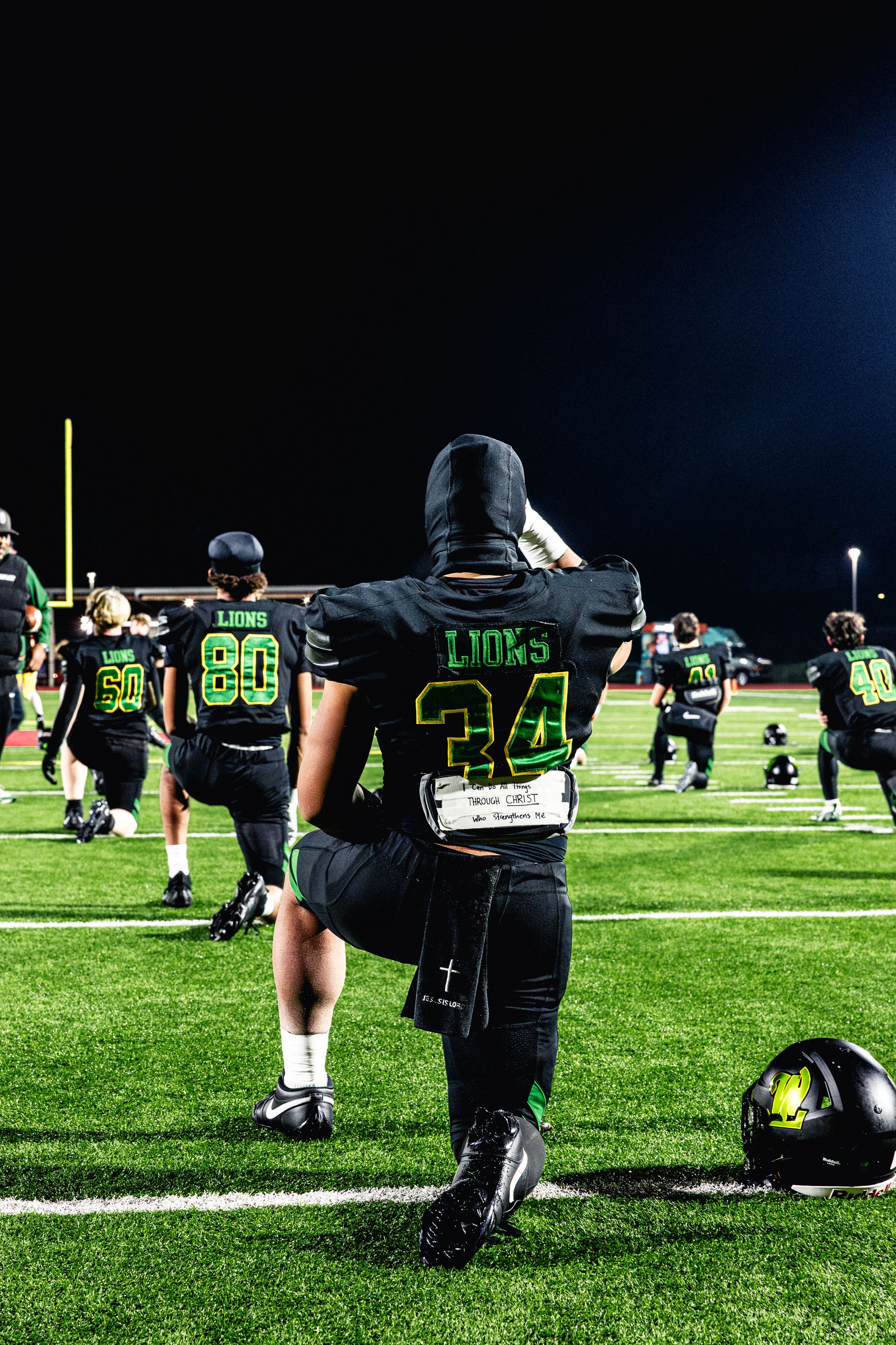 Football players in black uniforms with neon green numbers stretch on a field during nighttime practice.