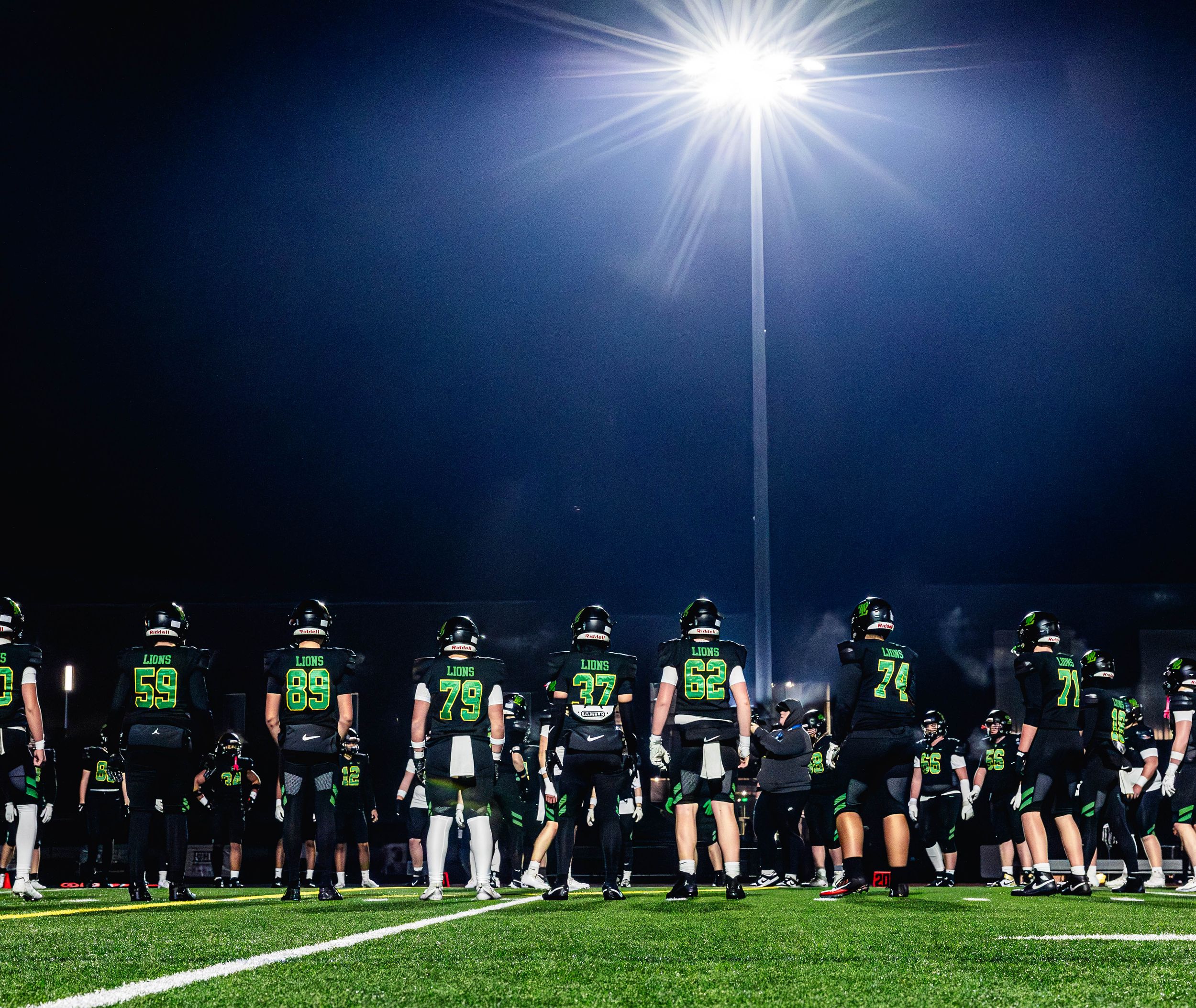 Soccer players line up under bright stadium lights at night during a training session on an outdoor field.