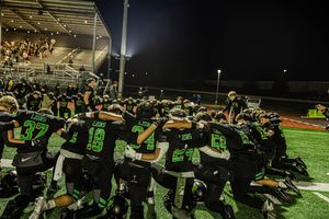 High school football players celebrate on the sideline during a night game under bright stadium lights.
