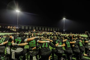 Football team huddles together in dark green uniforms under bright stadium lights at night.