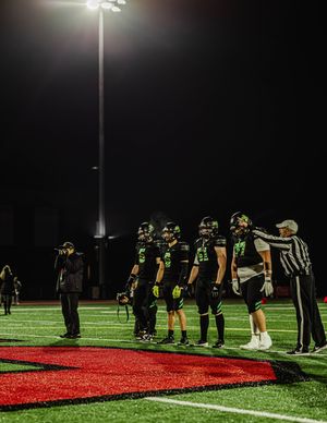 Football players and officials line up on red turf field under bright stadium lights at night.