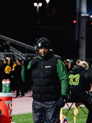 A person in a black puffer vest and green sweatshirt stands on a sports field sideline at night with stadium lights visible.