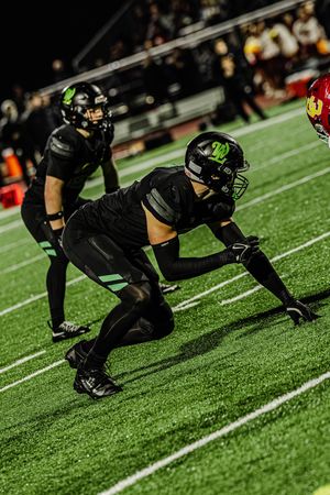 Football players in black uniforms and green helmets crouch in defensive stance on artificial turf.