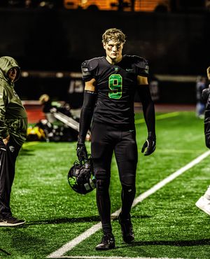 Players in black uniforms stand along the sideline of an illuminated football field at night.