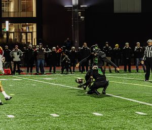 Football player kneels on artificial turf field during nighttime game with spectators and officials watching from sideline.