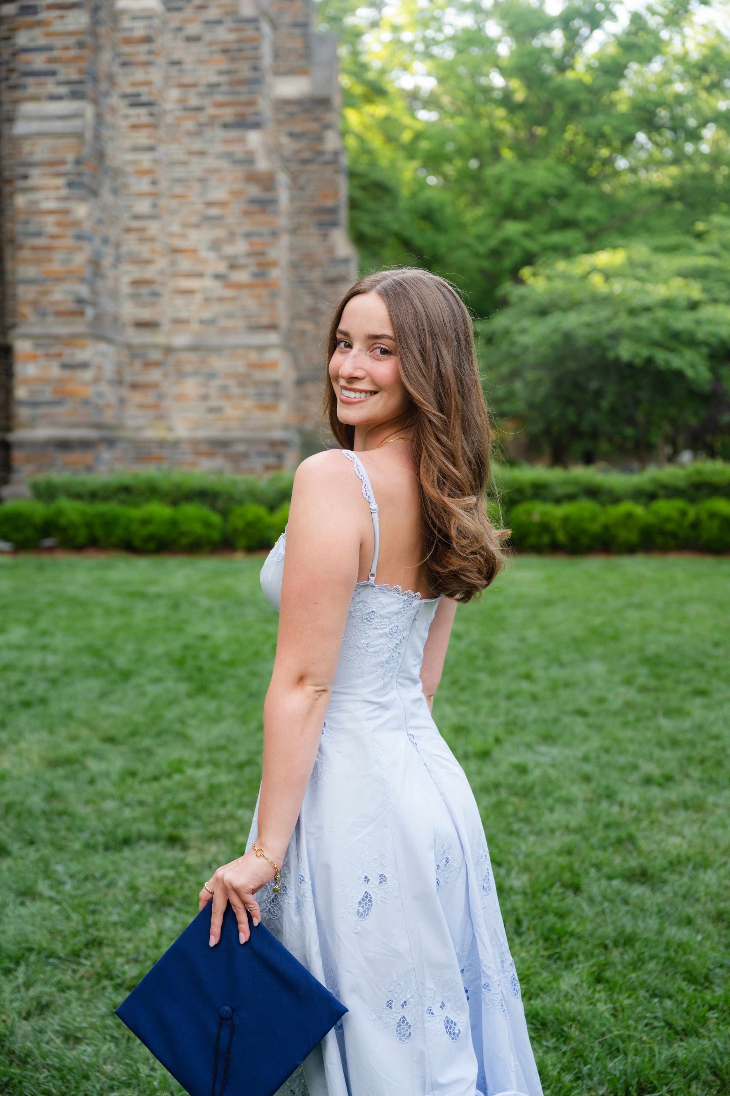 Smiling young woman in light blue gown holding navy graduation cap outdoors on green lawn near stone building.