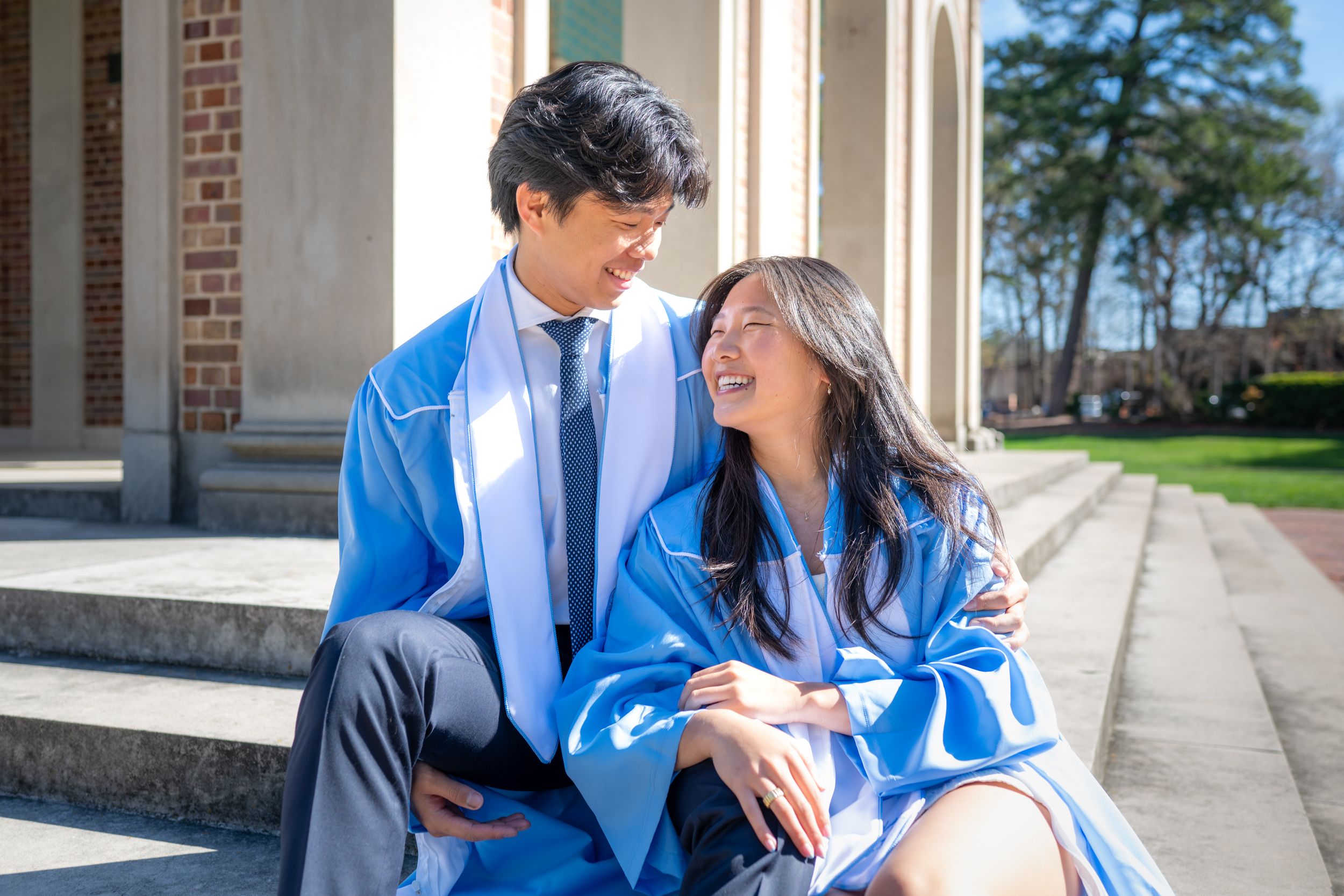 Two graduates in bright blue academic regalia share a joyful moment while sitting on stone steps outside.