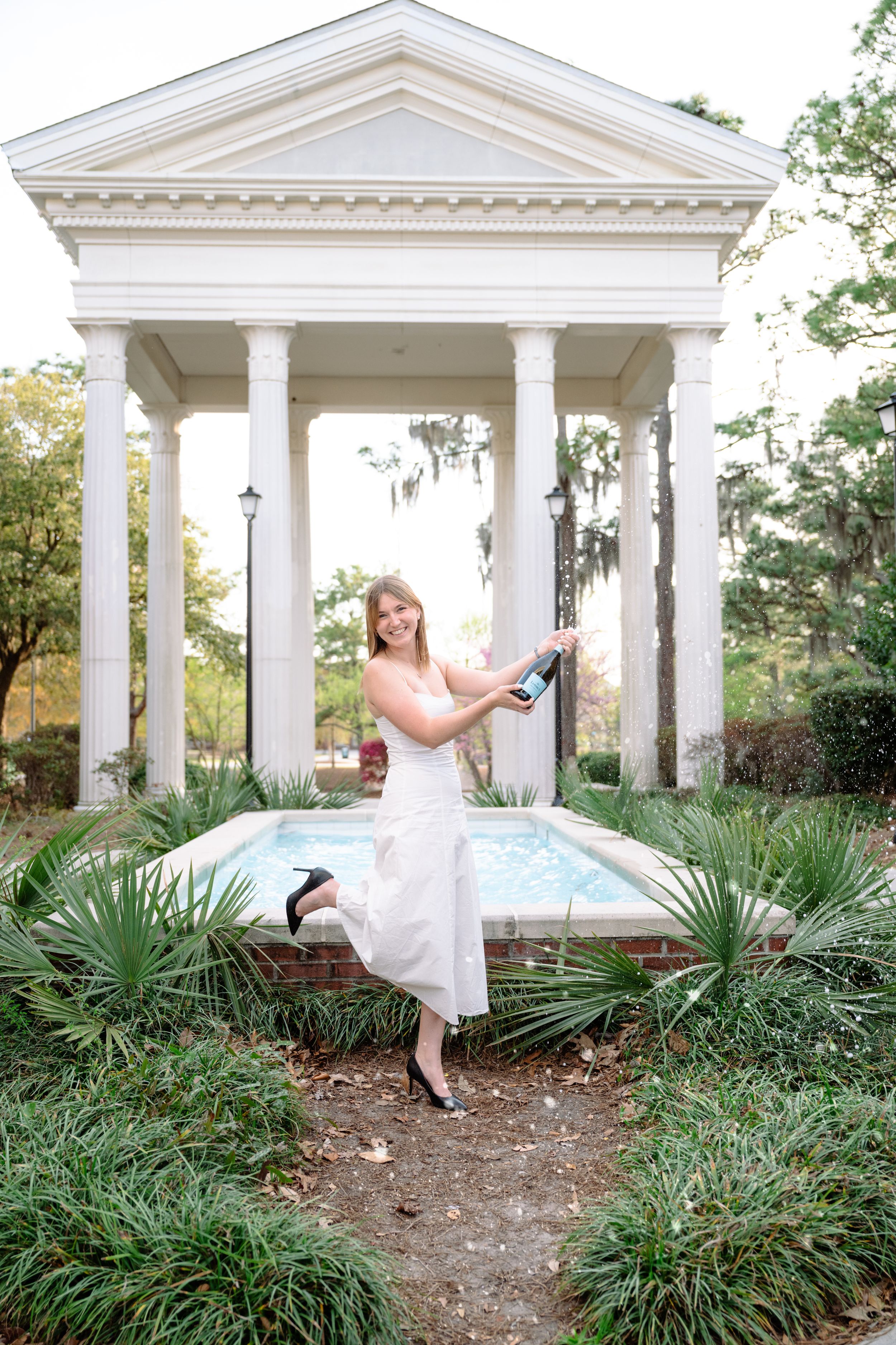 Smiling bride in white dress popping champagne bottle at elegant white gazebo garden fountain.