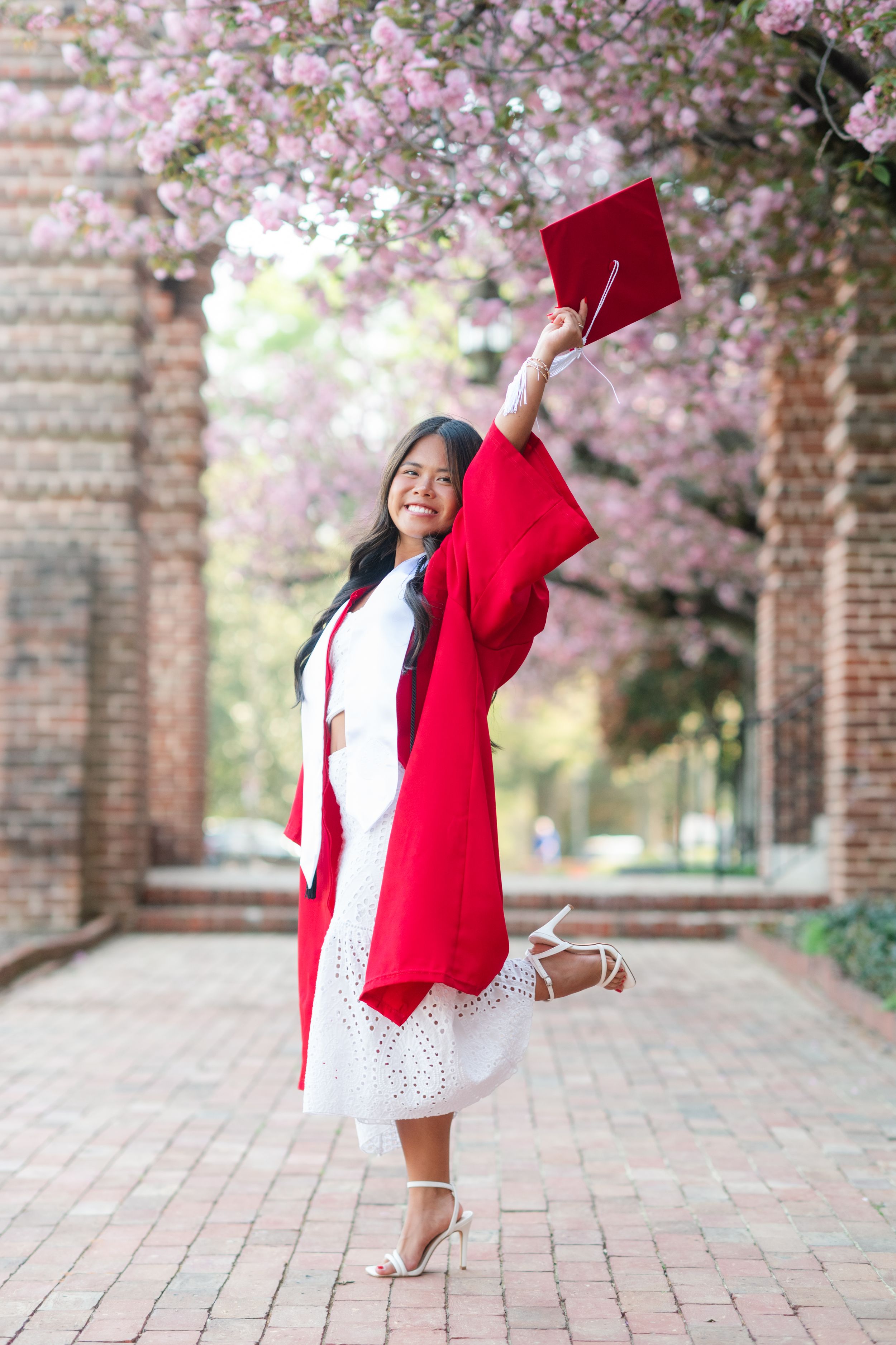 A joyful graduate in red gown raises cap under pink cherry blossoms on a brick pathway.