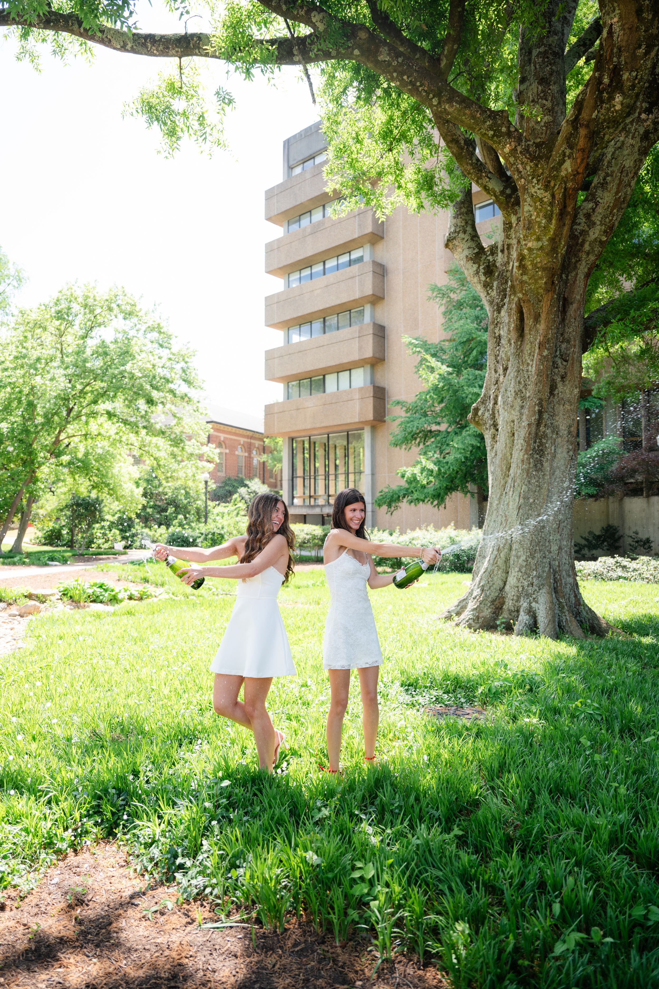 Two women in white dresses celebrate outdoors, spraying champagne near a large tree on a sunny day.
