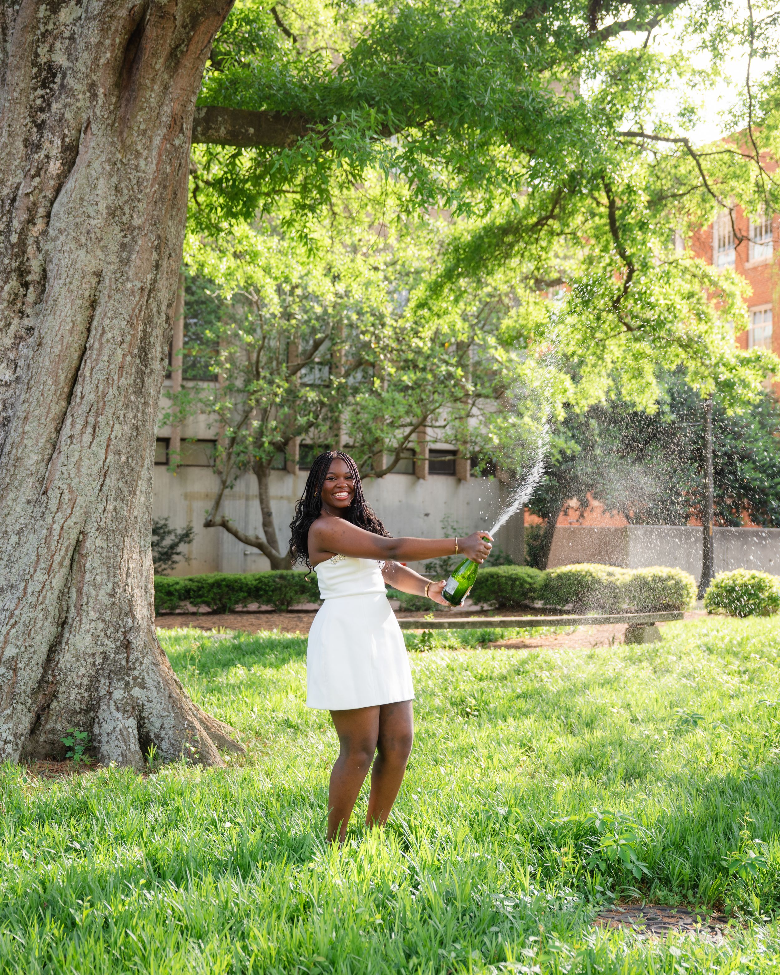 Young Black woman in white dress joyfully spraying champagne outdoors under a large tree on green grass.