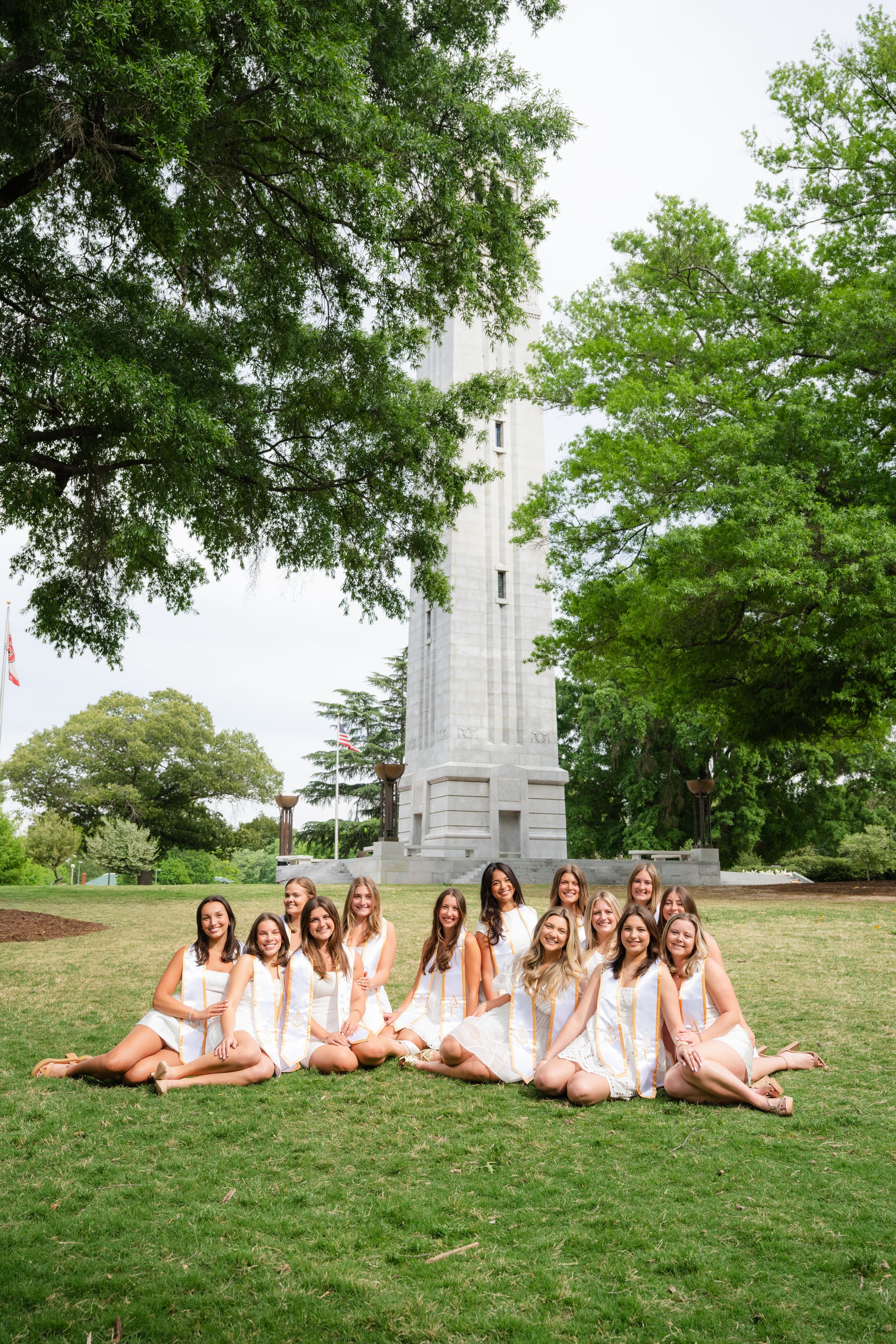 Group of women in white dresses sitting on grass in front of a tall monument surrounded by trees.