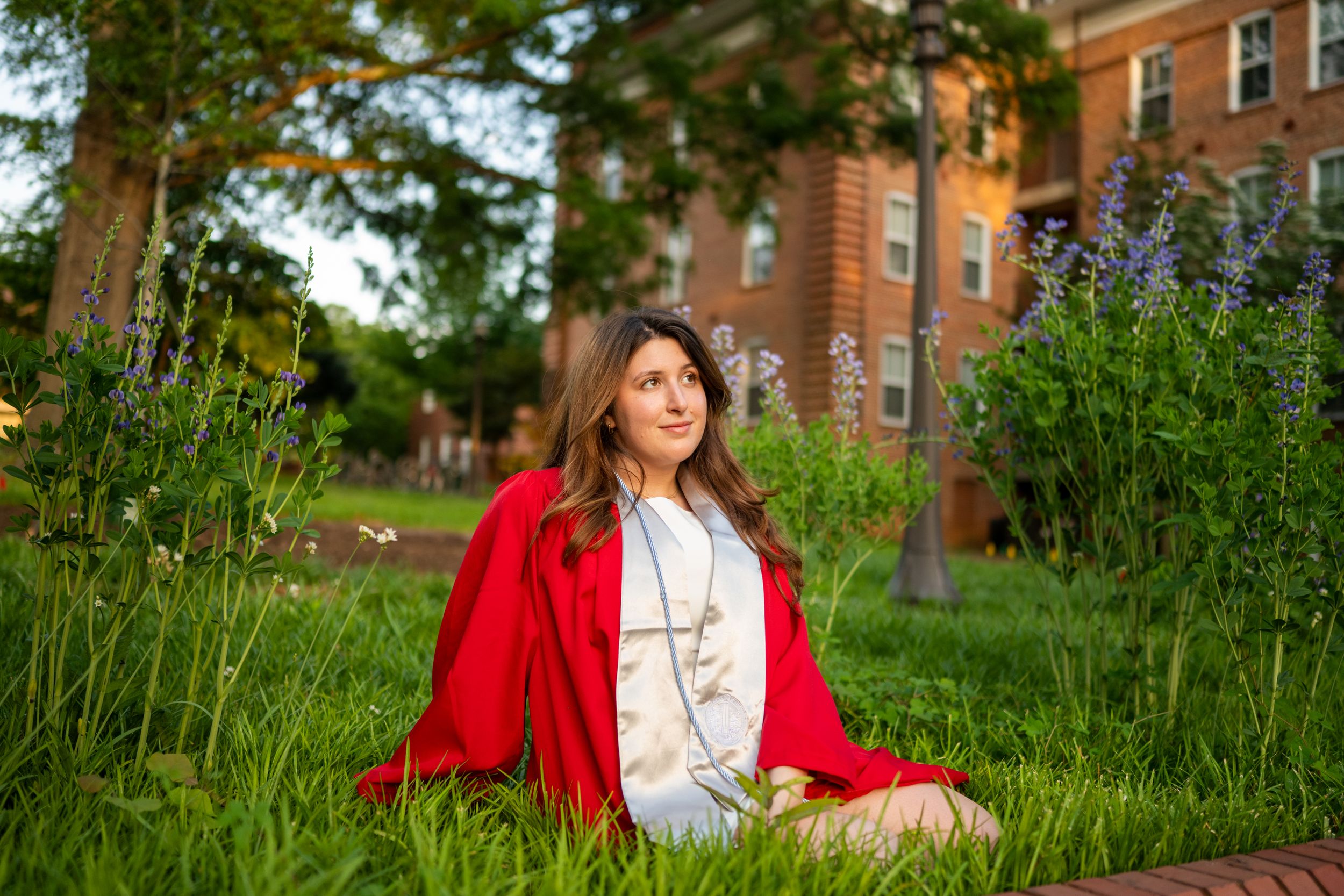 Person in a bright red jacket sits in a lush green garden setting with brick buildings in the background.