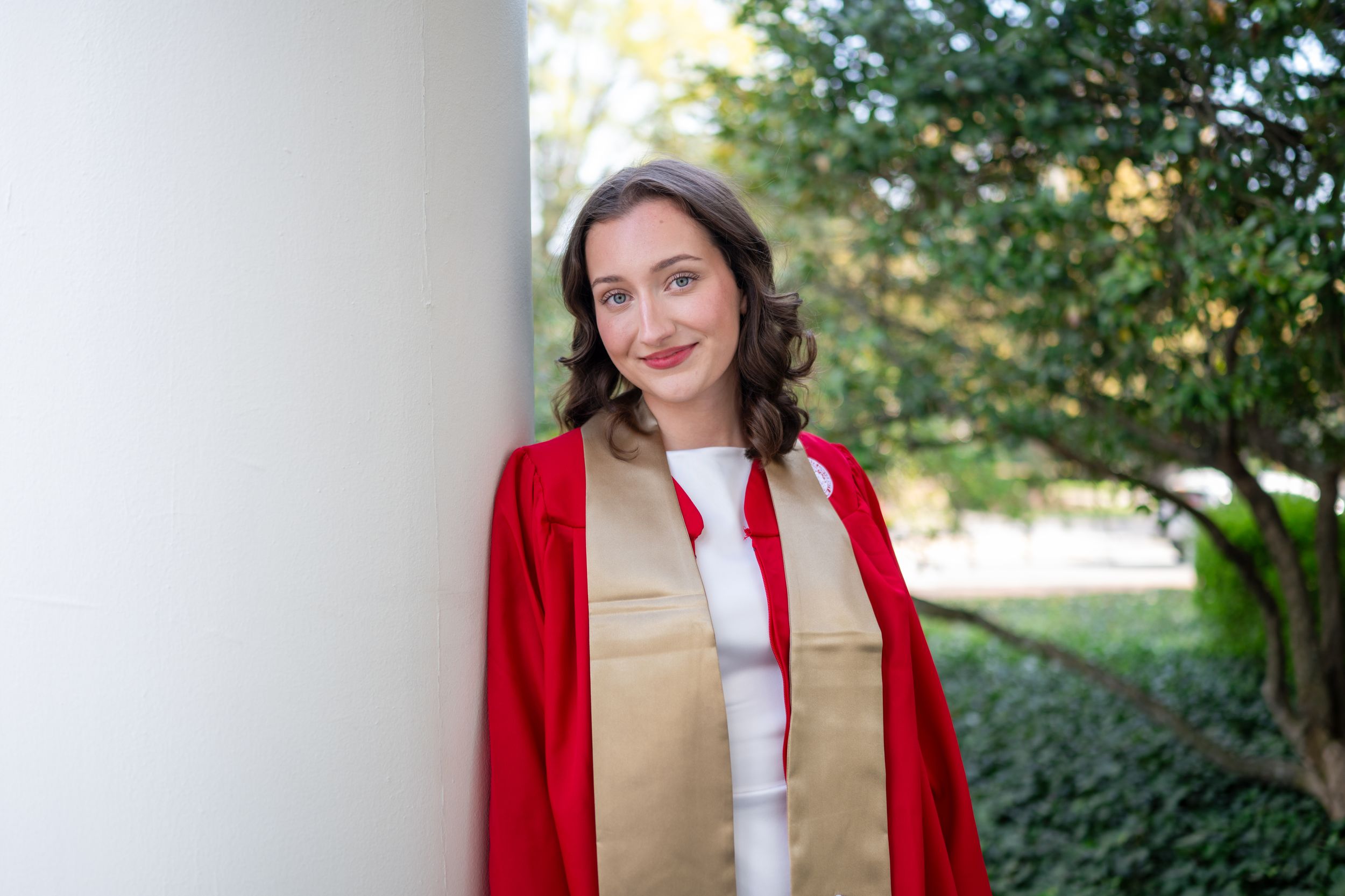 A person in a red blazer and tan vest poses outdoors near green foliage in a professional photo series.