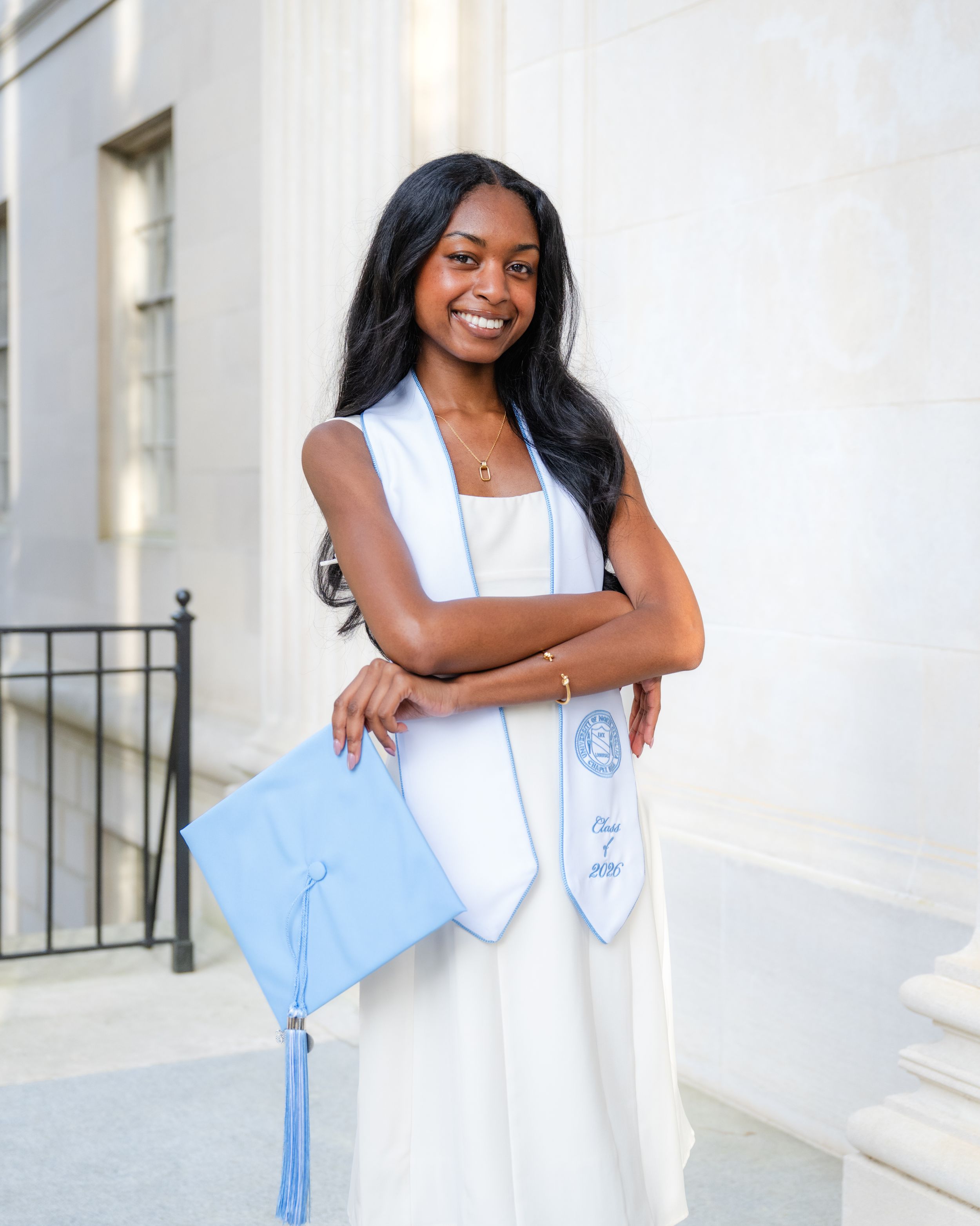 Young woman in white graduation gown holding light blue cap and stole, smiling outdoors at graduation ceremony.