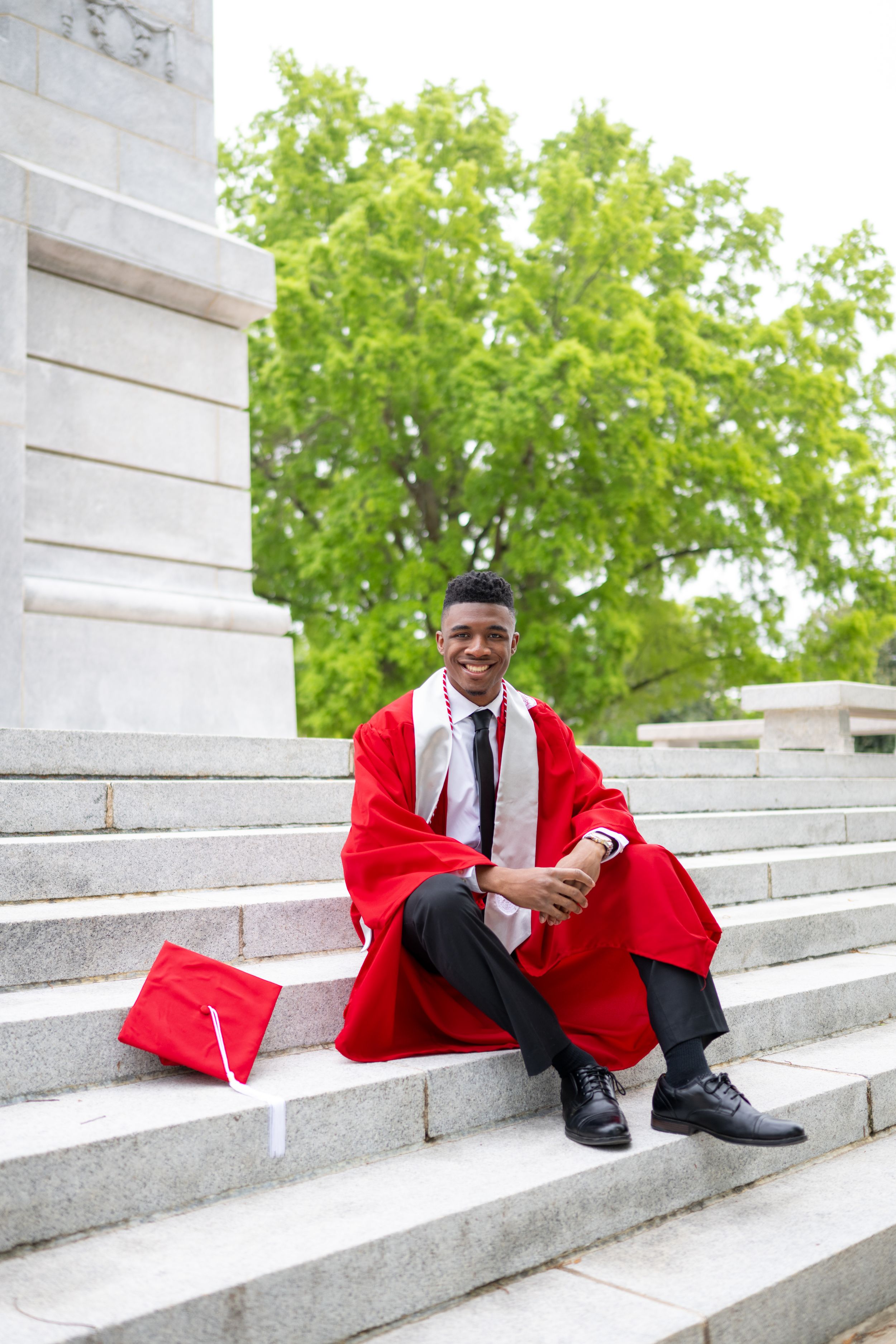 Young man in red graduation gown and cap sitting on stone steps, smiling proudly on his graduation day.