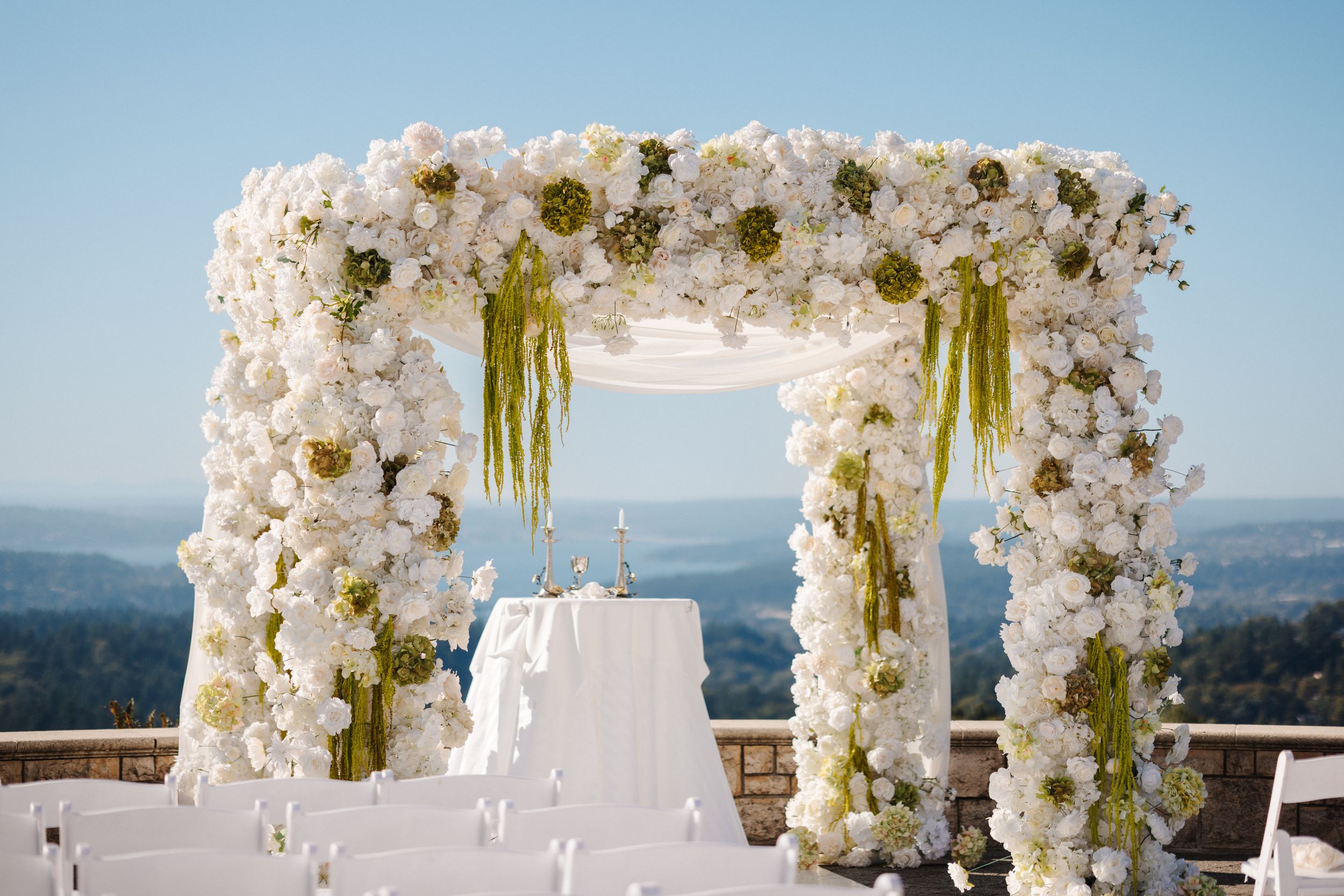 Elegant white floral arch with orchids and roses decorates an outdoor wedding ceremony overlooking a scenic mountain view.