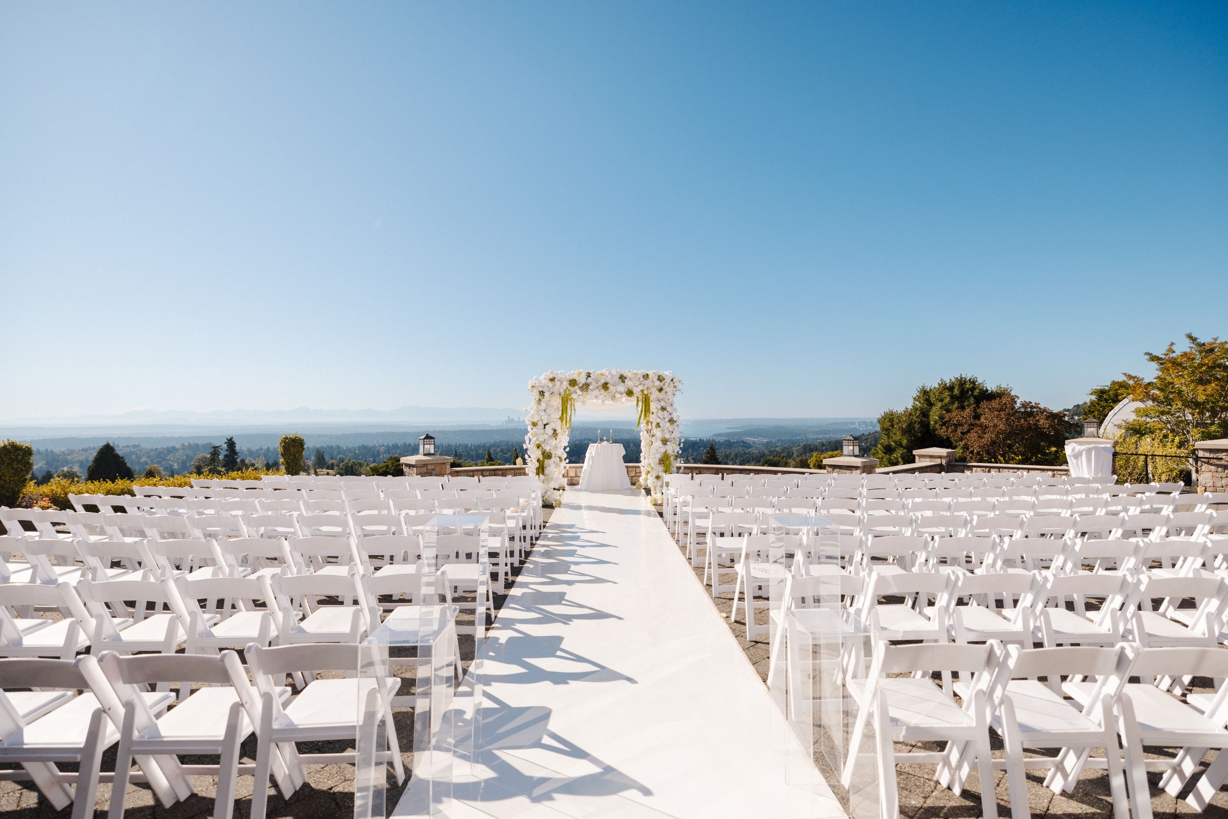 White chairs line up in rows facing a floral arch at an oceanfront wedding ceremony setup on a sunny day.