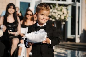 Ring bearer in a gray suit walks down the aisle carrying a white pillow during an outdoor wedding ceremony.