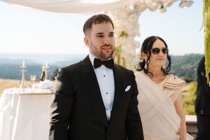 A couple in formal attire stands at an outdoor wedding ceremony with mountain views and decor in the background.