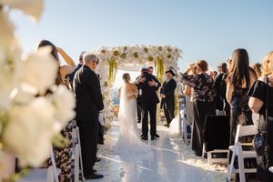 Wedding guests gather outdoors at a scenic venue during a sunset ceremony with white floral decorations.