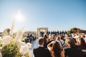 Outdoor wedding ceremony on a sunny day with guests seated and floral decorations against a bright blue sky.