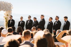 Wedding ceremony takes place outdoors with groomsmen lined up at the altar against a bright sky backdrop.