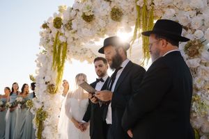A traditional Jewish wedding ceremony takes place under an elegant floral chuppah with white blossoms at sunset.