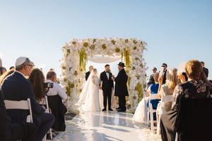 Wedding guests gather around a white floral arch during an outdoor ceremony at sunset.