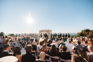 A panoramic view of wedding guests seated outdoors at a ceremony with blue skies and scenic backdrop.
