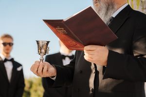 A person in formal attire holds a ceremonial cup while reading from a book during an outdoor ceremony.