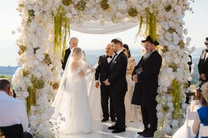 Wedding ceremony under a white floral arch with guests watching as couple exchanges vows at sunset.