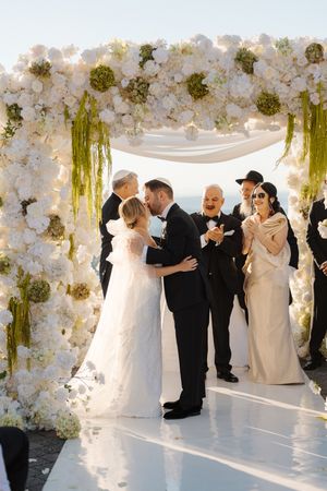 A romantic wedding ceremony takes place under an elegant white floral arch with cascading greenery and soft lighting.