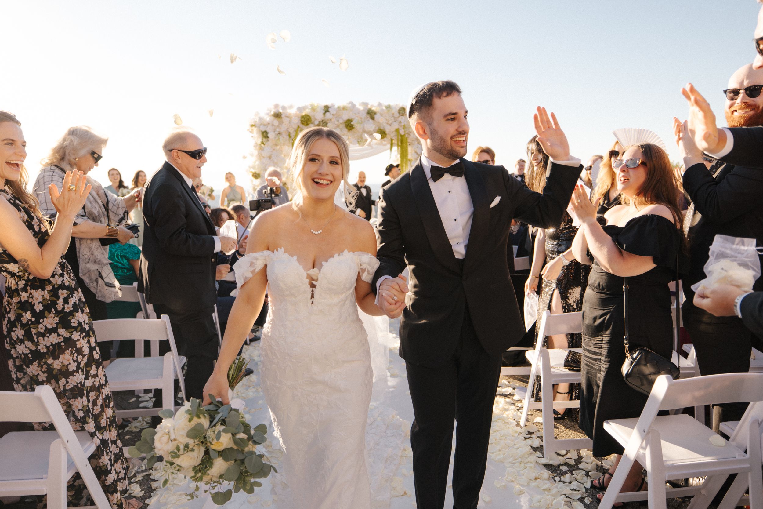 Newlyweds walk down aisle at outdoor ceremony while guests celebrate with white chairs and floral arrangements.