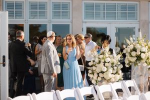 A row of empty white chairs arranged for an outdoor wedding ceremony with floral decorations and guests in the background.