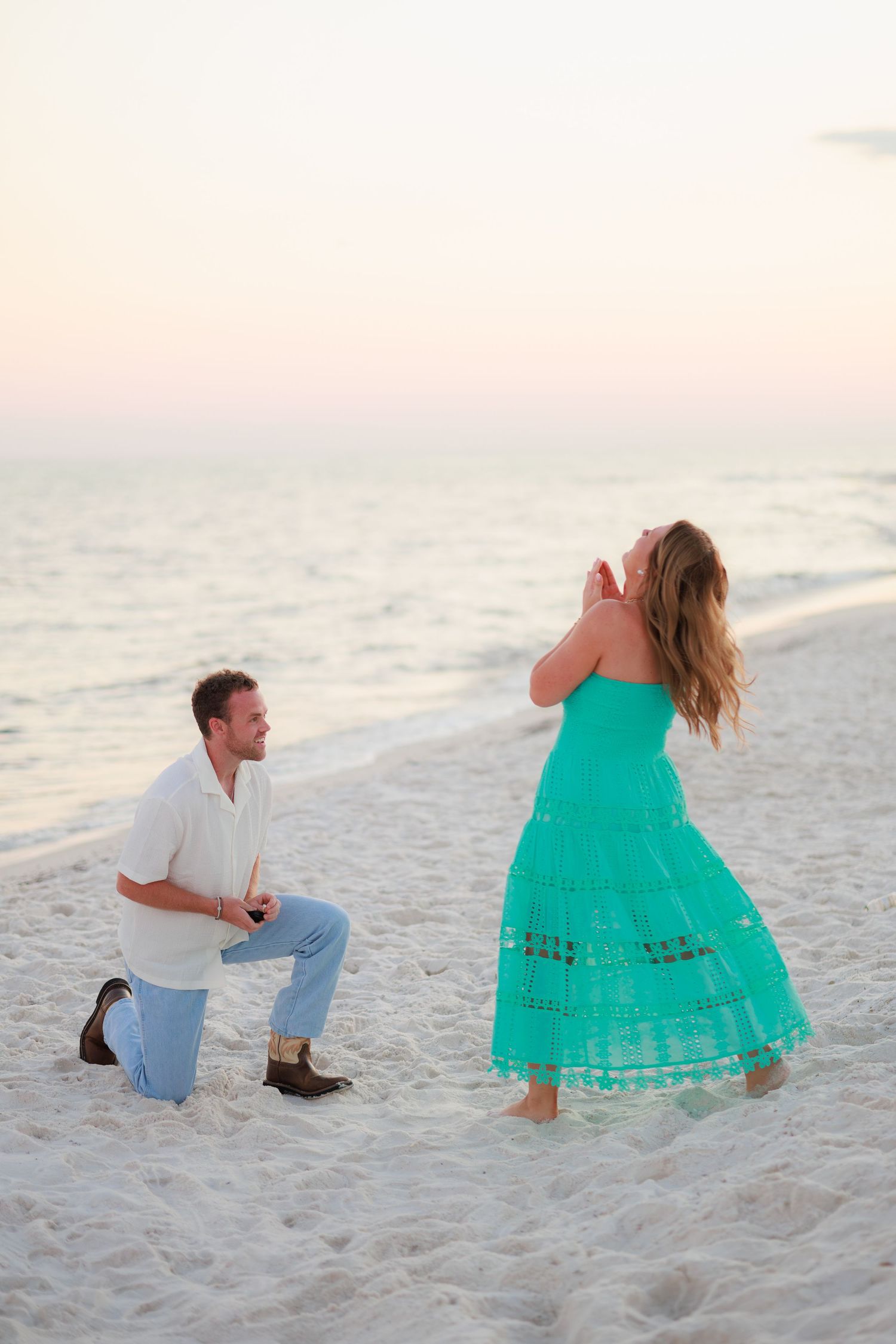 A marriage proposal scene on a sandy beach at sunset with someone in a turquoise dress and another person kneeling.