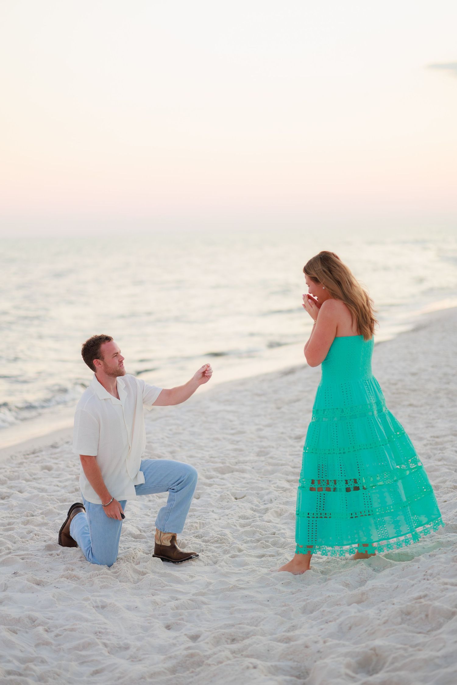 A couple in white and turquoise outfits poses playfully on a white sandy beach at sunset.