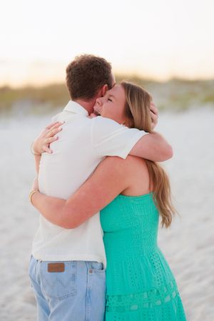 A couple embraces on a sandy beach at sunset wearing a white shirt and turquoise dress.