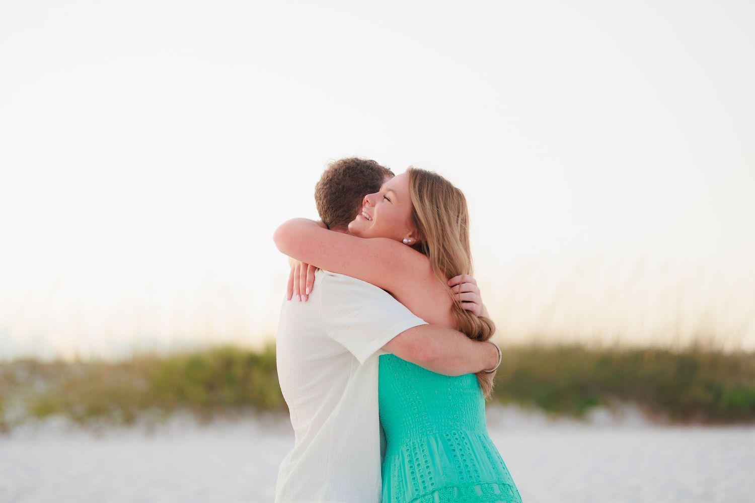 A couple embraces on a sandy beach during sunset while wearing white and turquoise outfits.