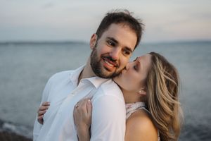 A romantic couple embraces on a beach at sunset wearing white outfits for their engagement or wedding photos.