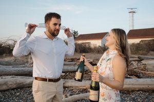 A couple celebrates with champagne on a beach at sunset with driftwood and a lighthouse in the background.