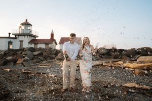 Couple stands together at sunset near a lighthouse on a rocky coastal cliff overlooking the ocean.
