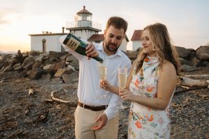 Couple celebrates with champagne at sunset near a lighthouse on rocky coastal cliffs.