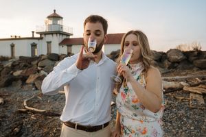 A couple enjoys champagne together near a lighthouse on rocky coastal terrain at sunset.
