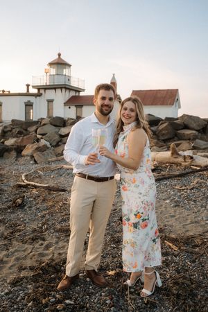 A couple poses with champagne glasses in front of West Point Lighthouse on a rocky beach at sunset.