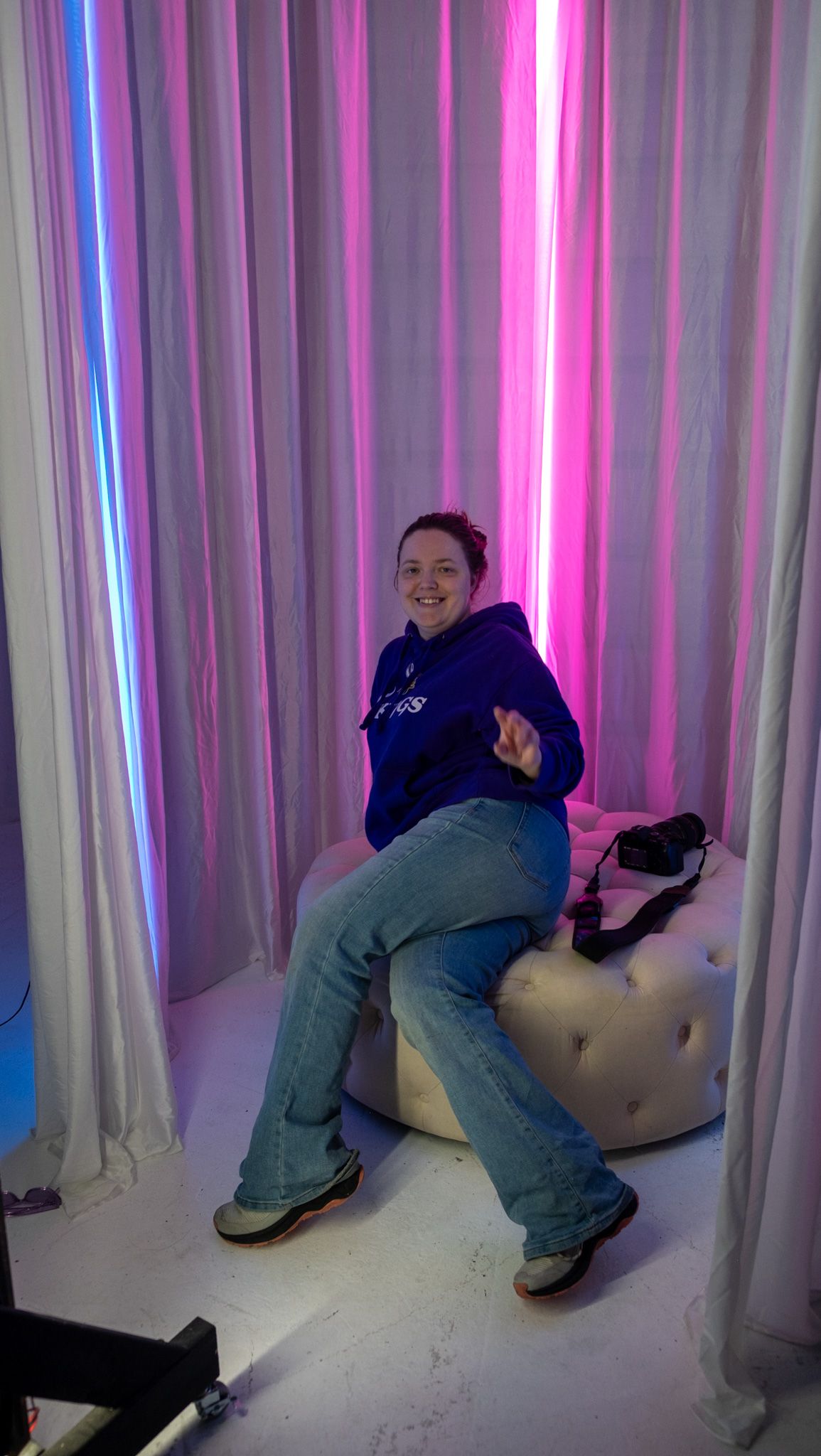 Behind the scenes photo showing colorful pink and blue lighting setup with white curtains and a person lounging on a couch.