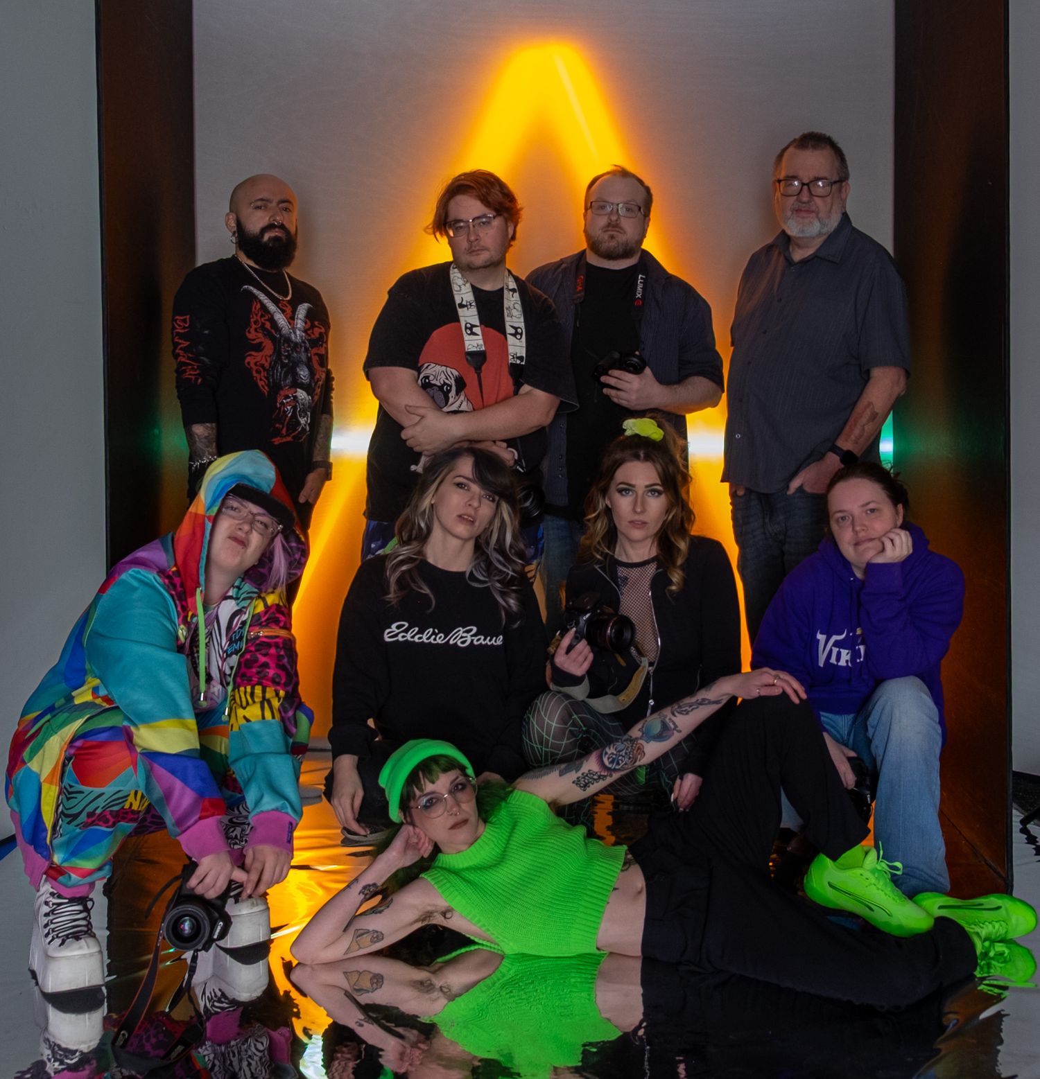 A group poses in front of a yellow-lit wall wearing colorful outfits including neon accessories and casual streetwear.