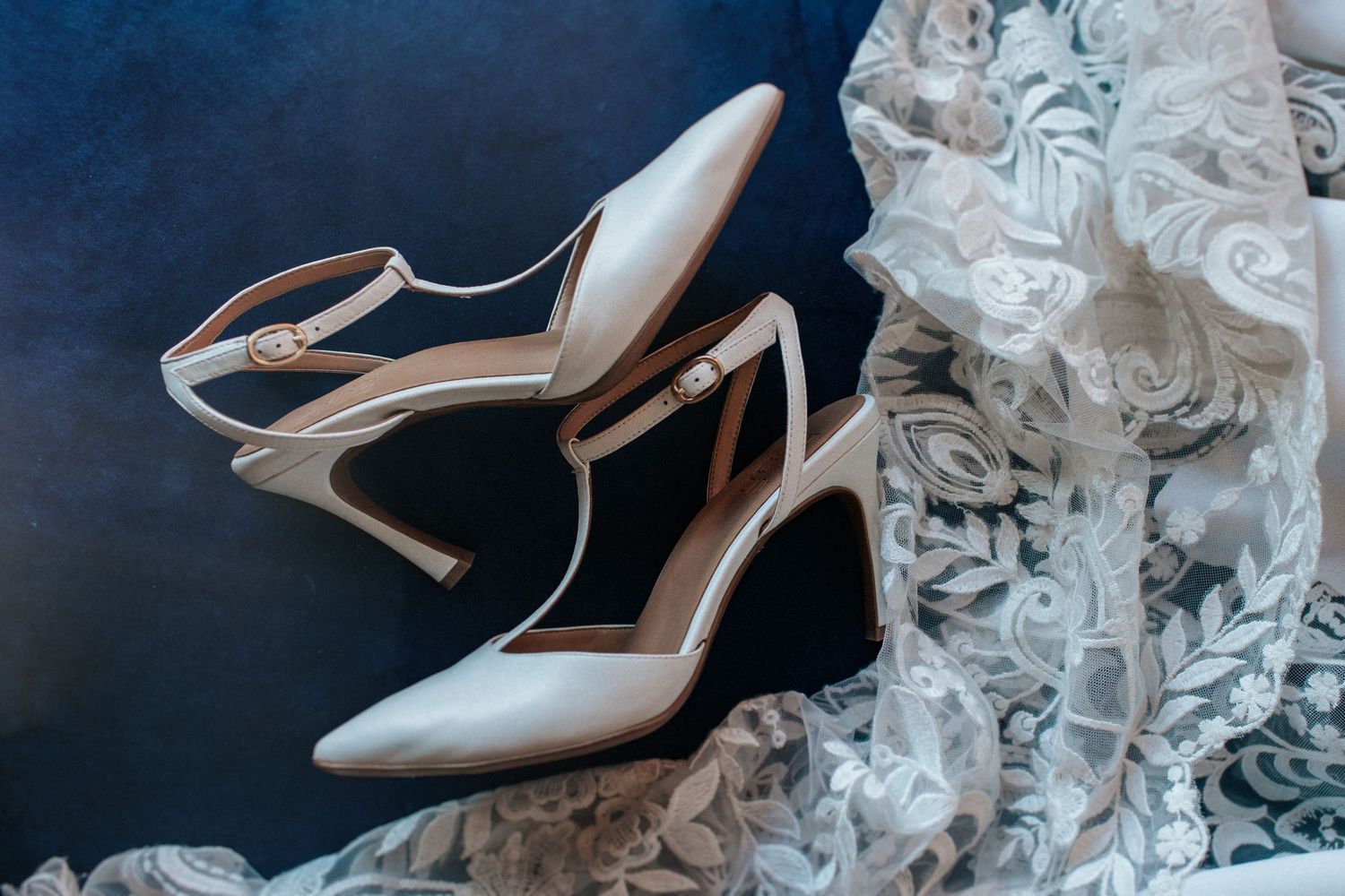 Elegant bridal shoes and lace details arranged on a dark blue background.
