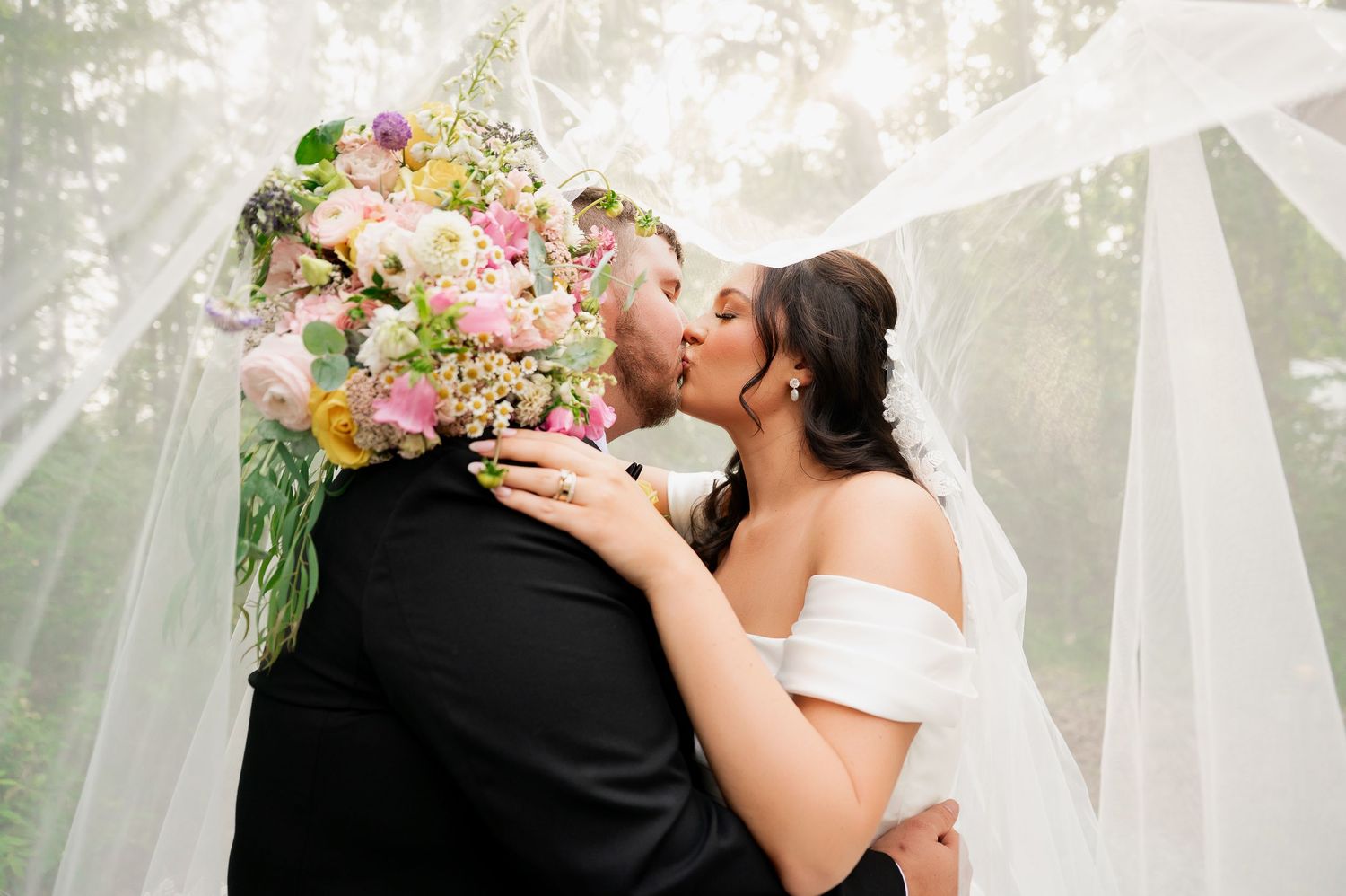 Romantic wedding kiss under white draping with colorful floral bouquet.